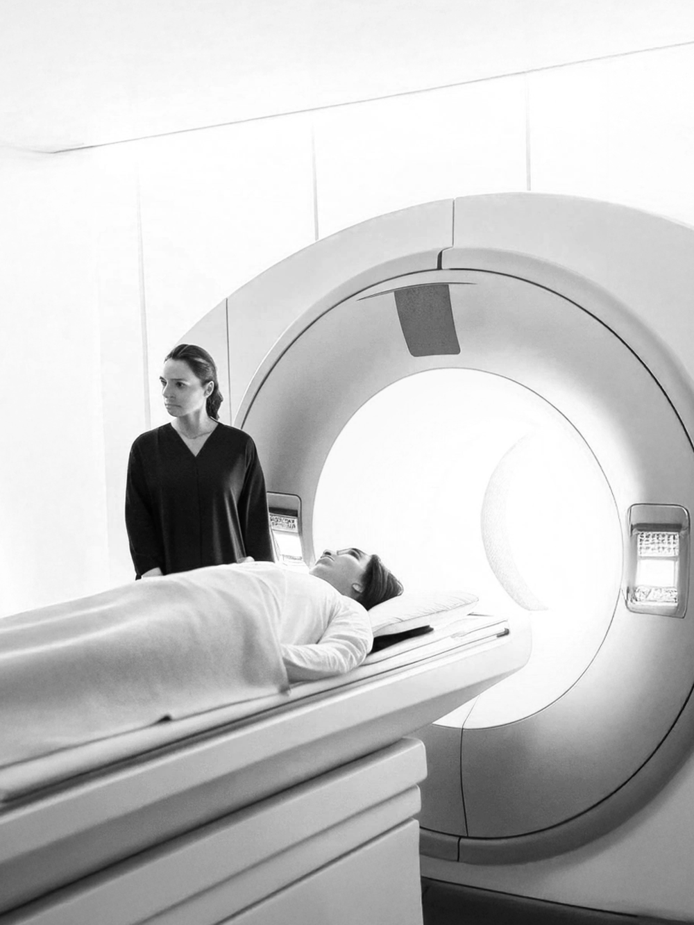 A woman lying on a hospital bed inside an MRI machine, with a female medical professional standing nearby in a clinical setting.