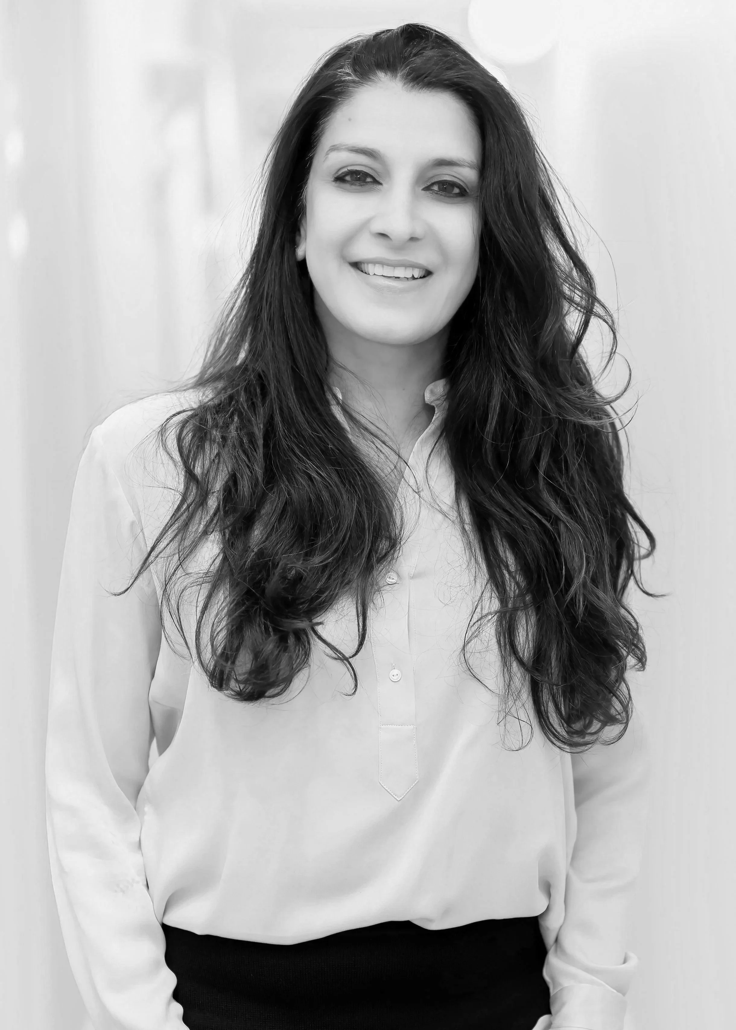 A black and white photo of a woman with long wavy hair, smiling and wearing a buttoned shirt.
