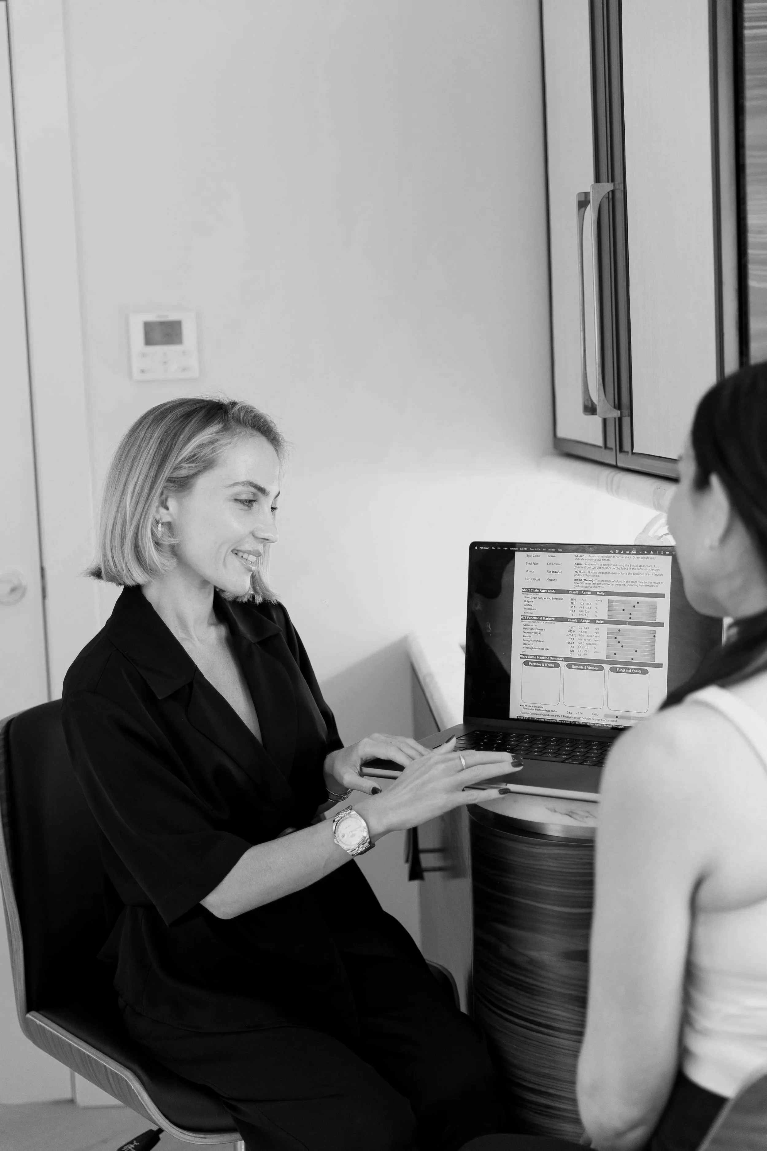 A woman sitting at a desk showing a smile to another woman with a laptop in front of them.
