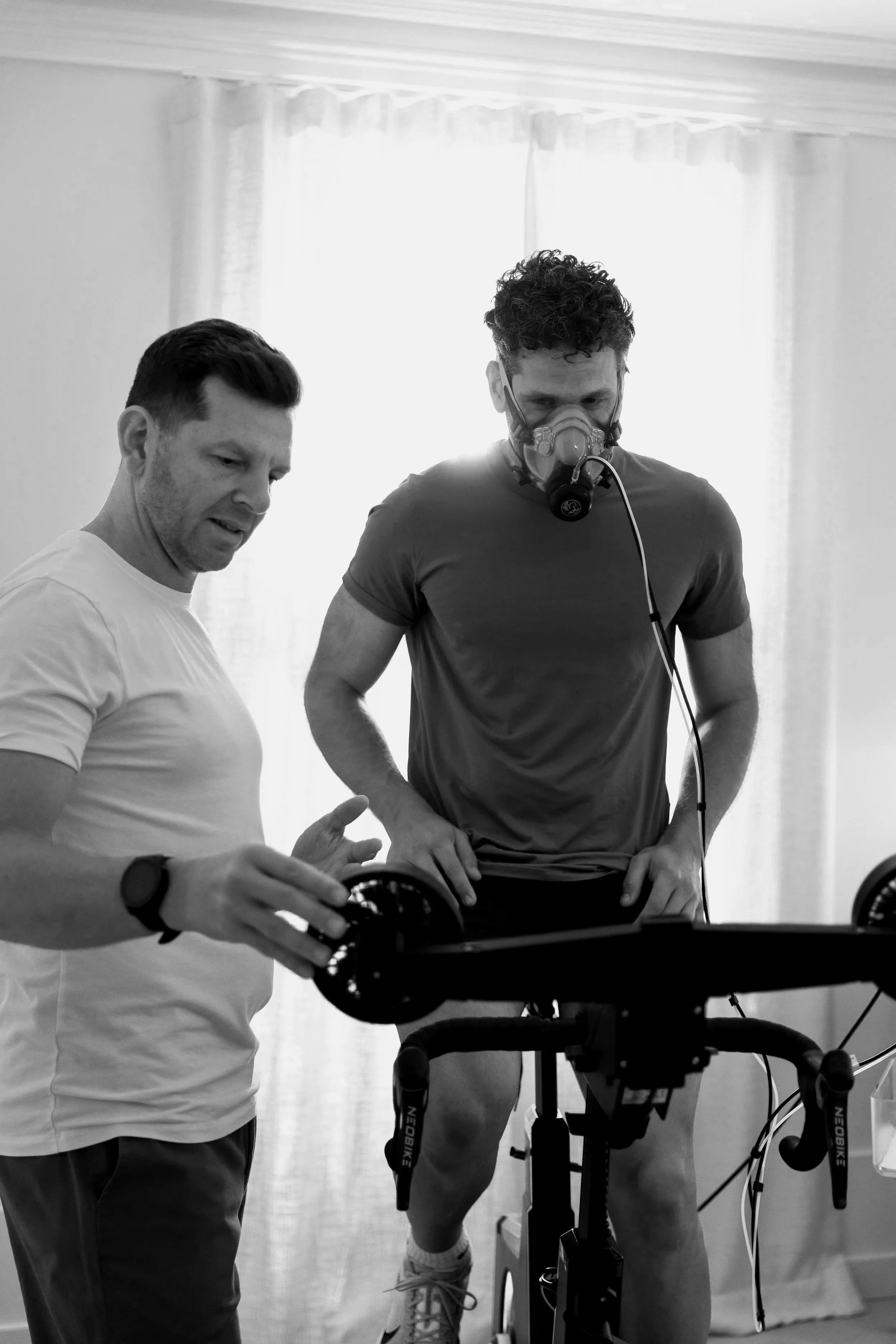 Two men observing a stationary bicycle, one wearing a mask connected to a device, and the other explaining or adjusting settings, indoors with sunlight coming through curtains.