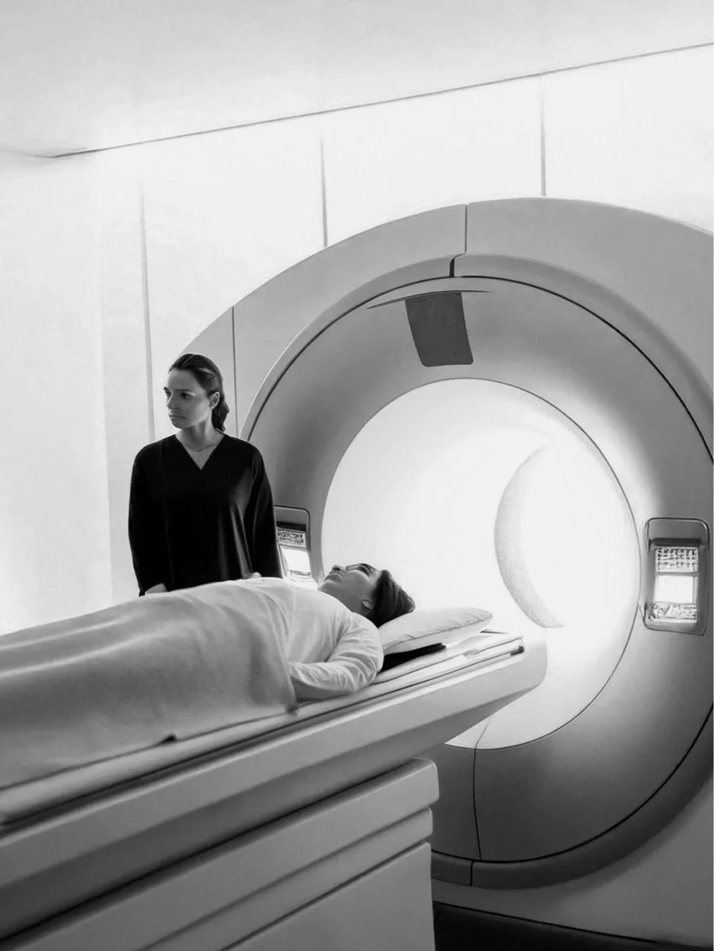 A woman lying on a hospital MRI or CT scan table with her eyes closed, and another woman standing next to her, in a medical imaging room with a large circular scanner.