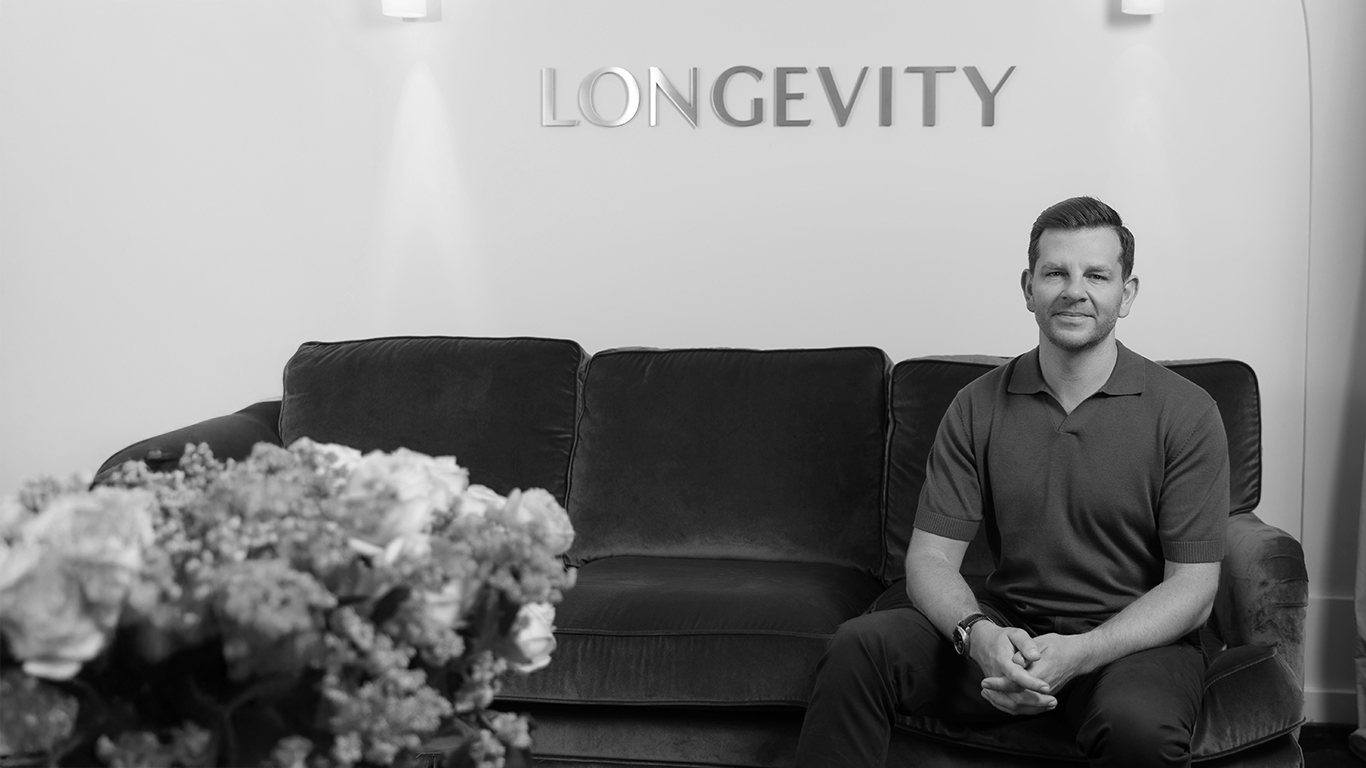 A man sitting on a couch in a room with the word 'LONGEVITY' on the wall behind him, and a vase of flowers in the foreground.