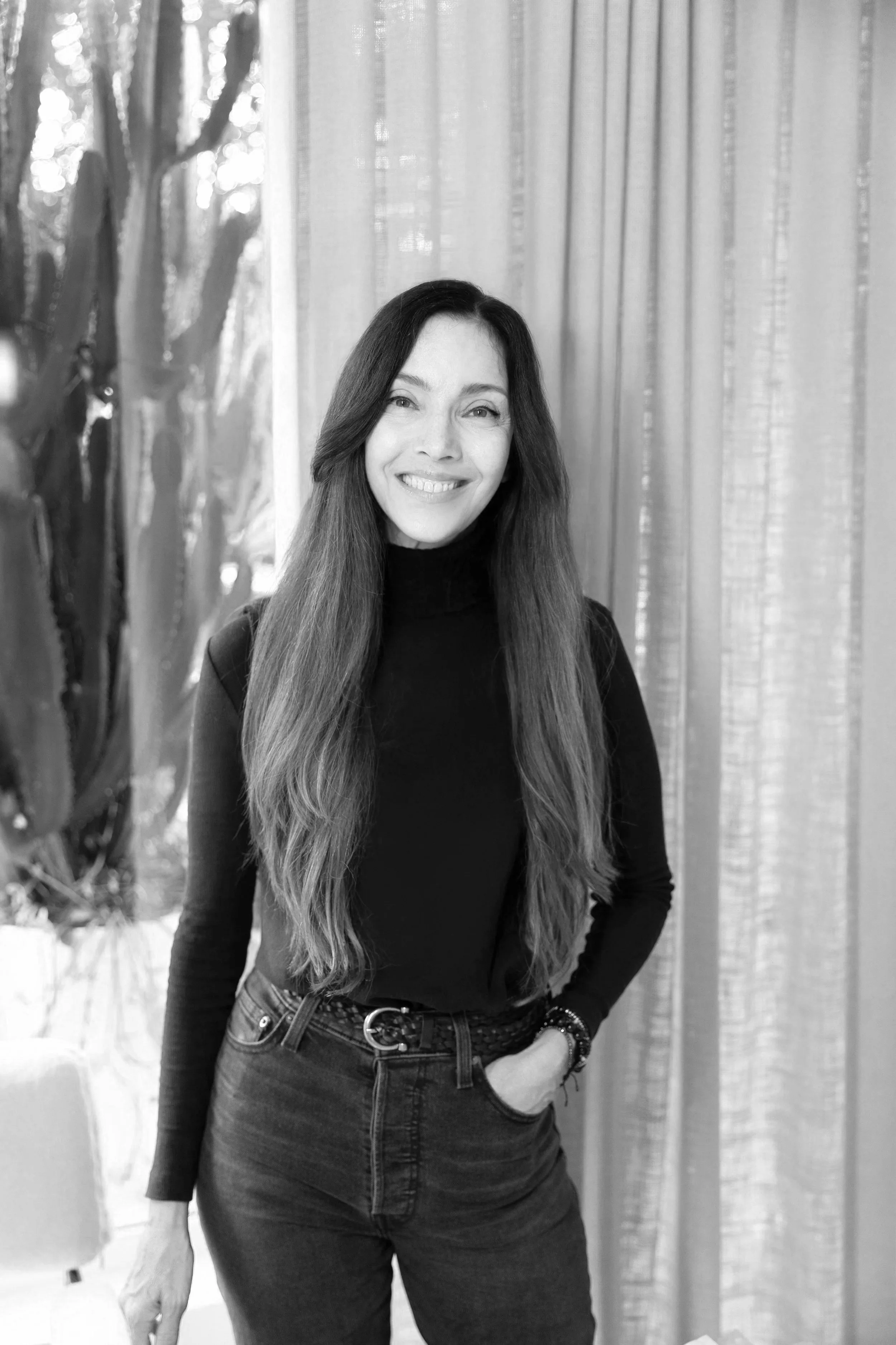 A smiling woman with long wavy hair, wearing a black turtleneck and dark jeans, standing indoors in front of a background with vertical striped curtains and a couch on the left side.