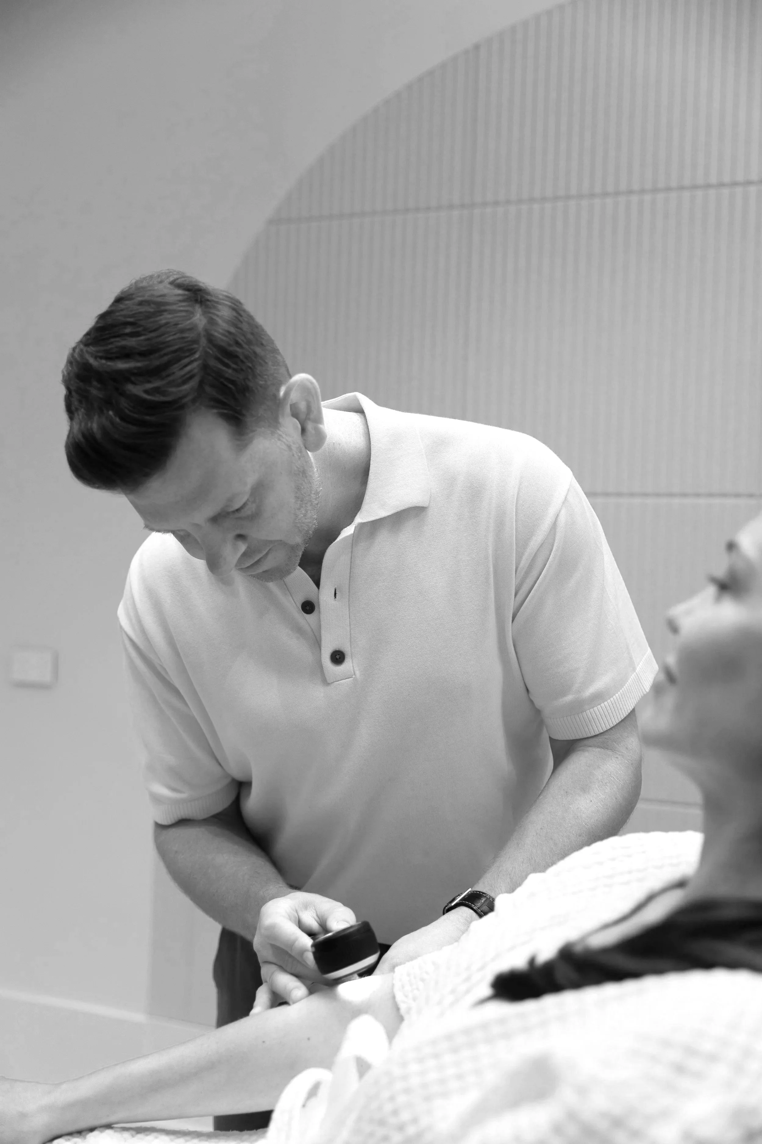A man in a white polo shirt using a handheld ultrasound device on a woman's arm in a medical setting.