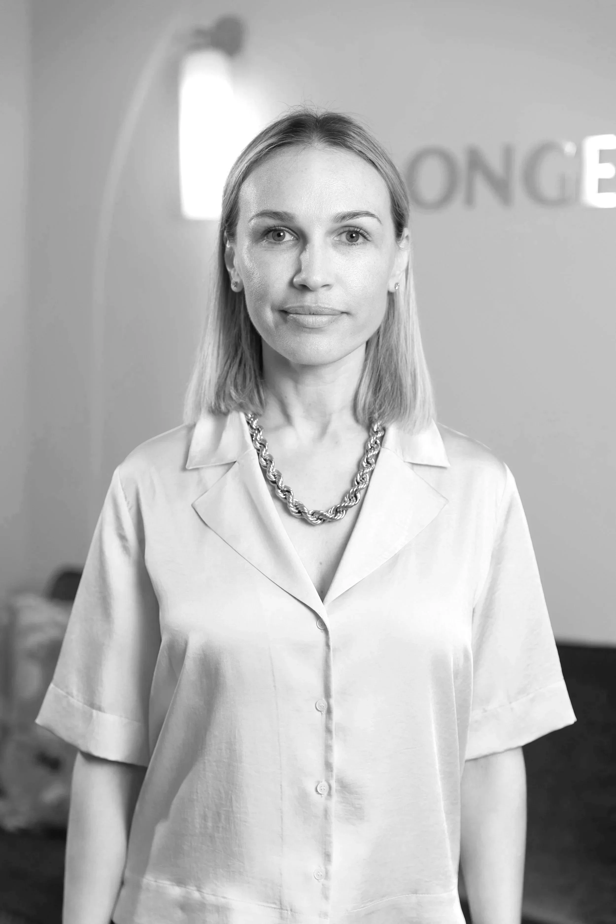 Black and white photo of a woman with shoulder-length hair, wearing a light-colored collared shirt with buttons and a chunky chain necklace, standing in front of a blurred background with the partial word 'LONGE' visible.