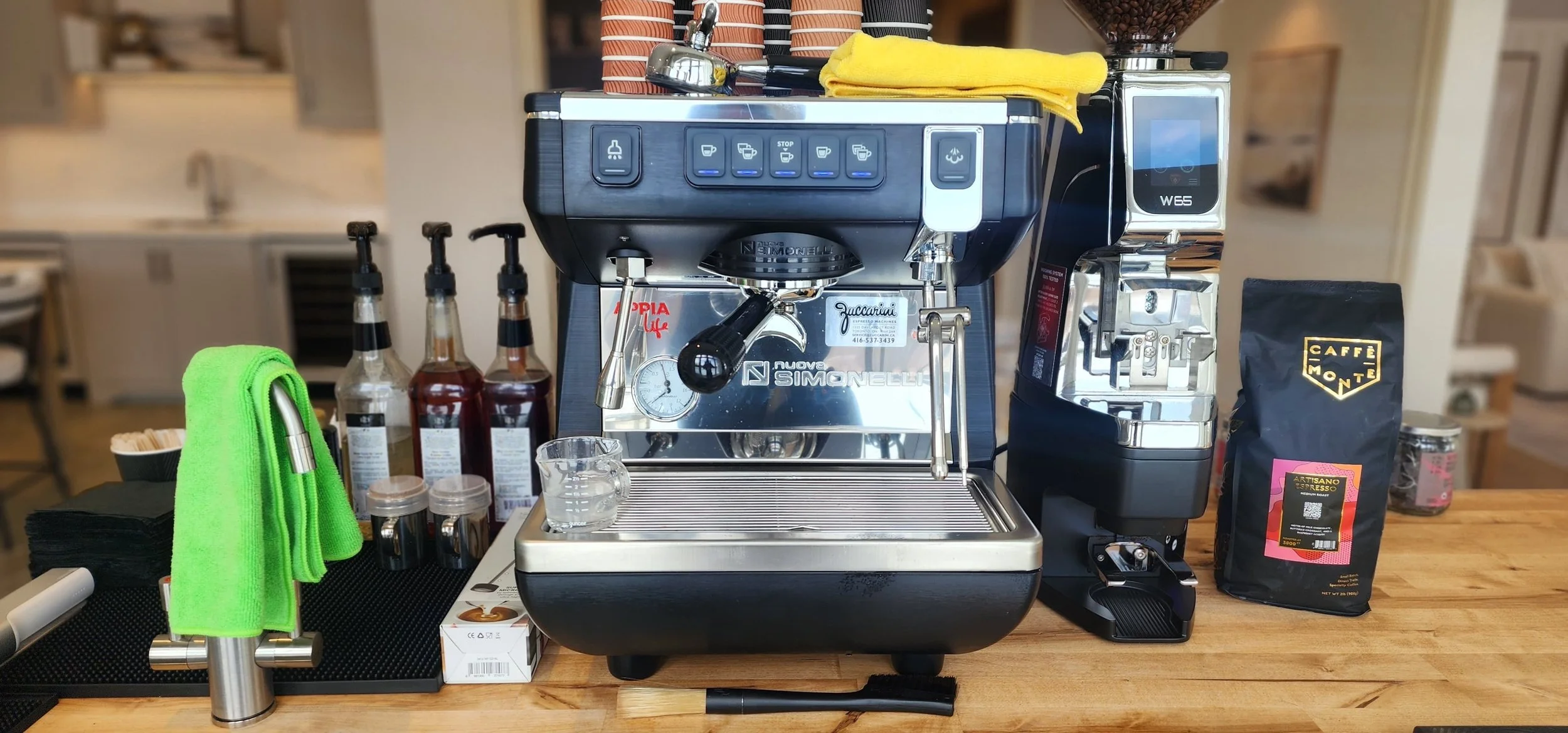 A coffee station featuring a professional espresso machine, a grinder, a roast coffee bag, three syrup bottles, a small glass of water, green cloth, black napkins, and small containers, all set on a wooden countertop in a cafe or kitchen setting.