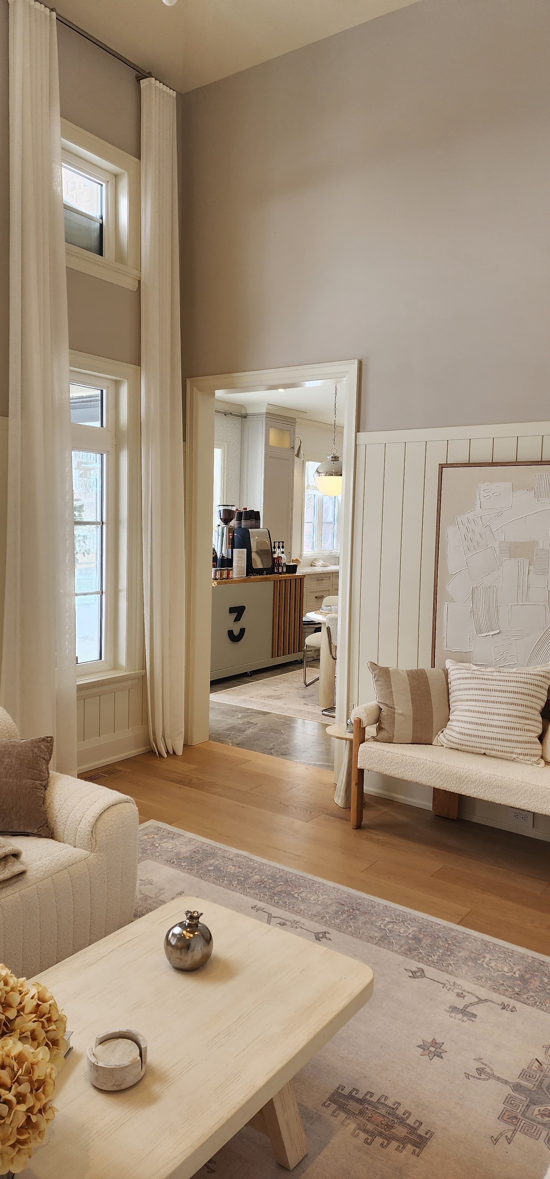 Living room with cream-colored sofa, patterned pillows, light wood coffee table with decor, area rug, large windows with white curtains, doorway to kitchen, light-colored walls, and high ceiling.
