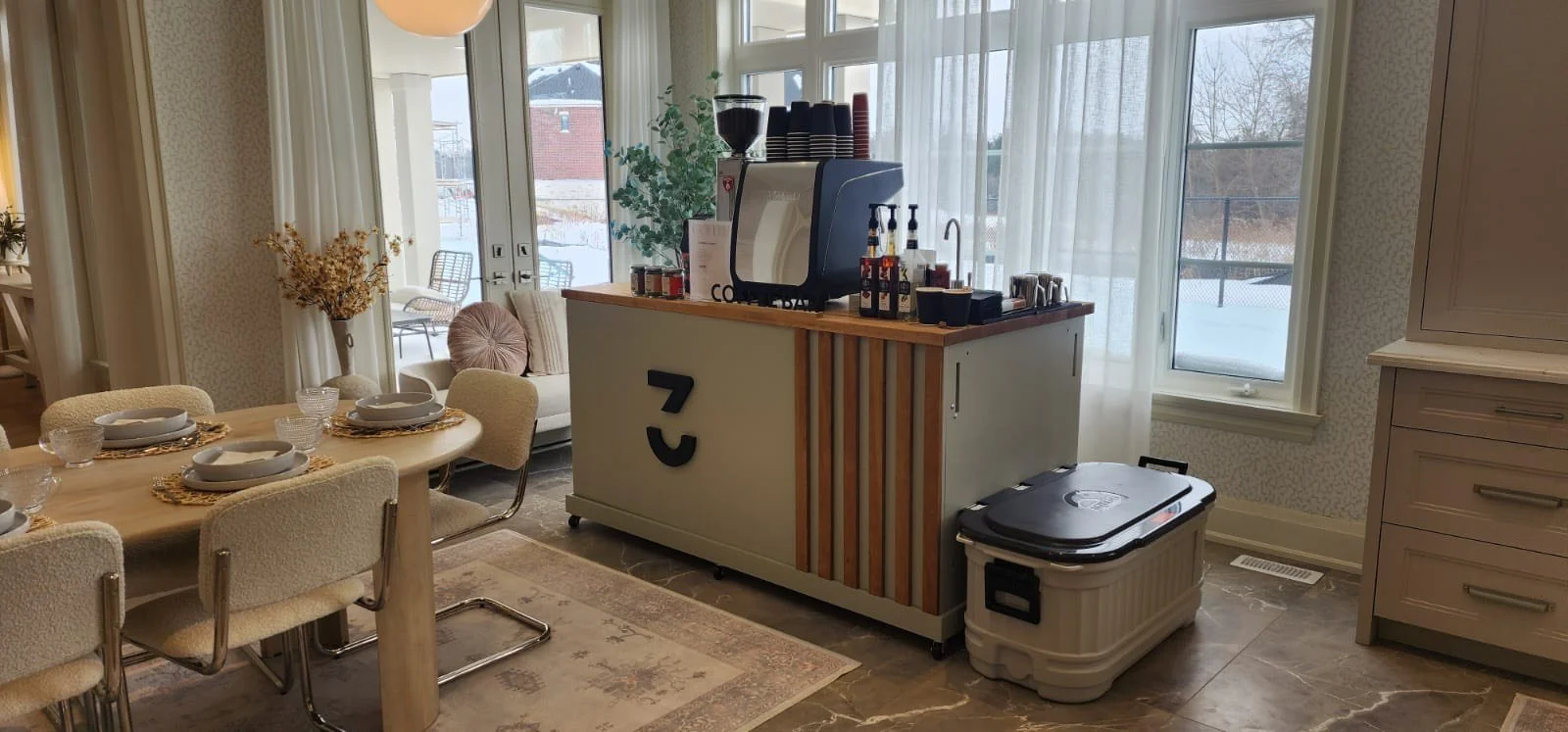 A dining area with a round table set for six, next to a light-colored sideboard with the number '7' on it, a small bar area with coffee cups, bottles, and a retro TV, and large windows with sheer curtains showing a snowy outdoor scene.