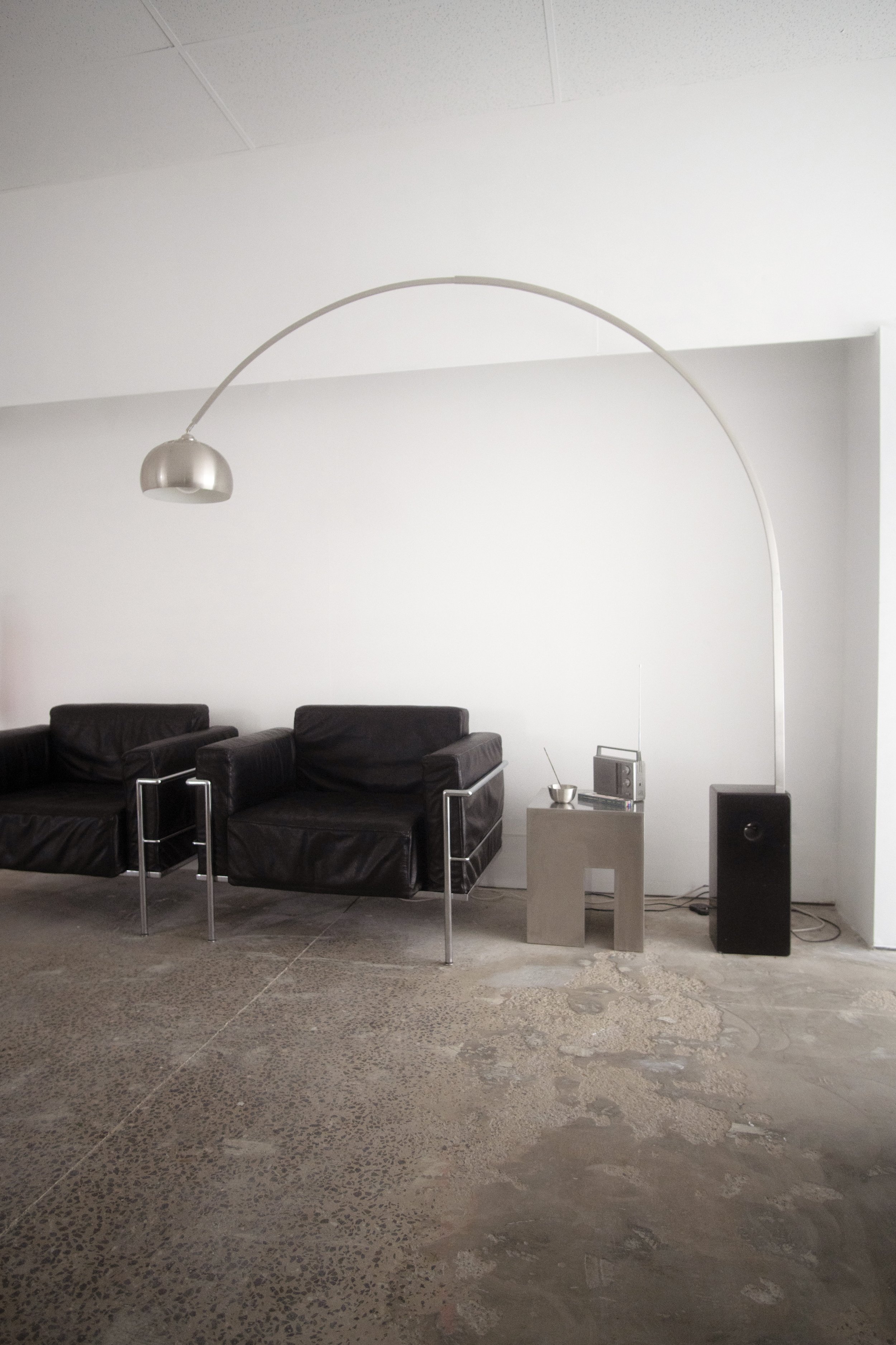 Minimalist living room with two black leather armchairs, a modern standing arc floor lamp, small side table with a bowl, radio, and speaker.