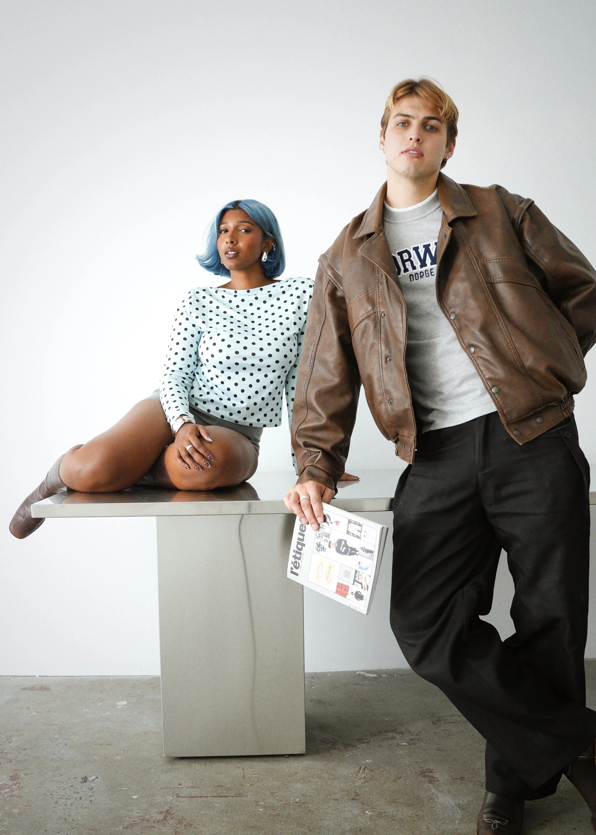 A young woman with blue hair and a young man with blond hair posing in front of a white wall. The woman is sitting on a gray table with one leg down and one knee up, wearing a polka dot top and beige boots. The man is standing, leaning on the table, wearing a brown leather jacket over a gray sweatshirt and holding a newspaper.