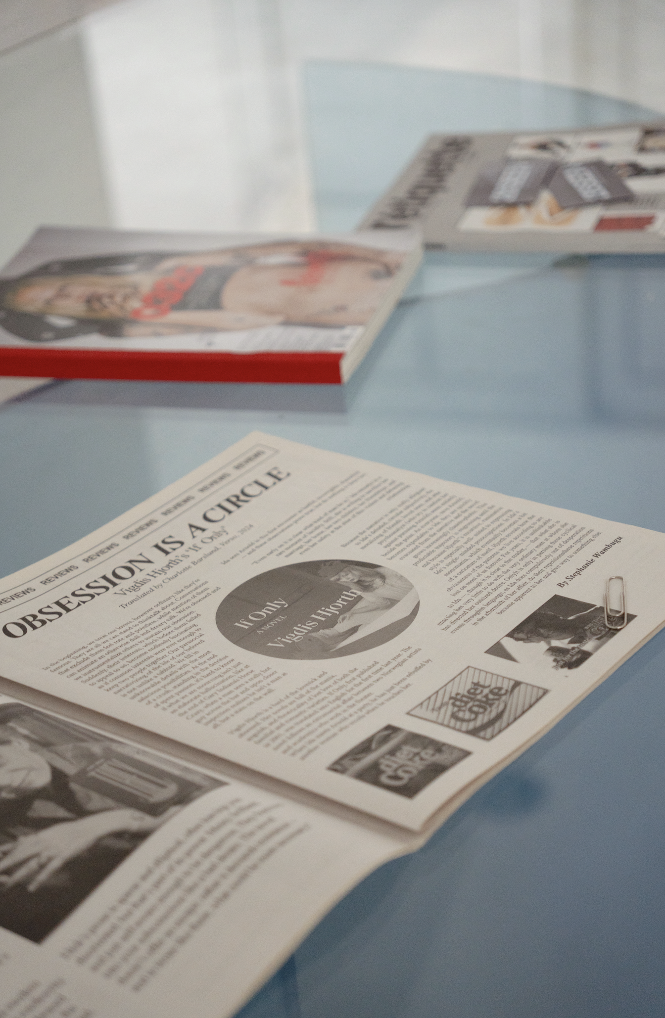 A newspaper titled 'Obsession Is a Circle' on a blue table, with a paperclip attached and a magazine in the background. The newspaper has a circular photo and text, with additional images and articles.