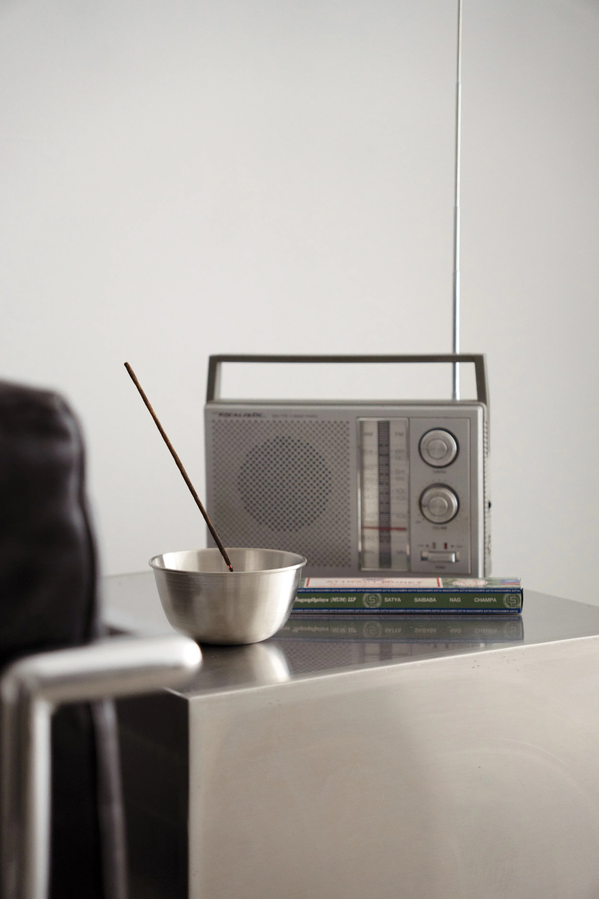 A stainless steel bowl with a matchstick inside, placed on a reflective surface with a vintage radio and a book in the background.