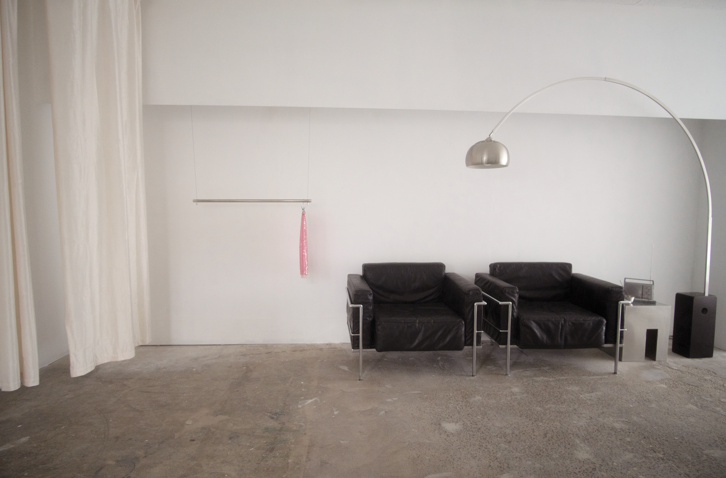 Minimalist living room with two black leather armchairs, a small side table, a floor lamp with an arched arm, a hanging pink cloth, and a remote on one of the tables. The room has bare concrete floor and plain white walls.