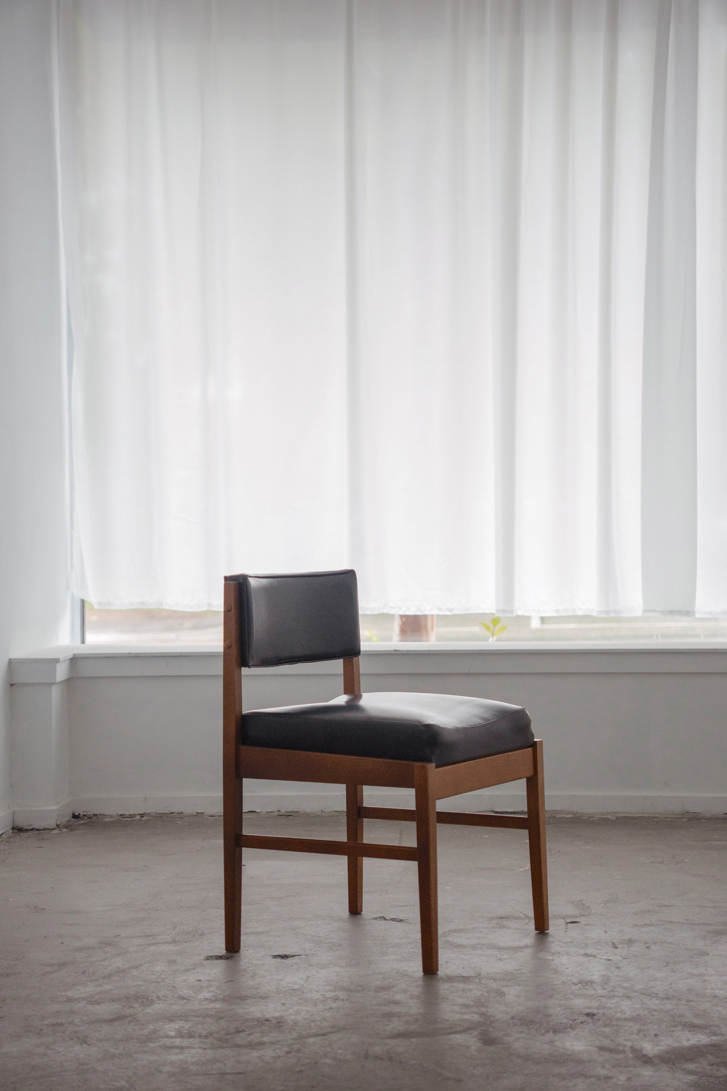 A black and brown wooden chair with a cushioned seat and backrest positioned in front of white curtains in a minimally decorated room.