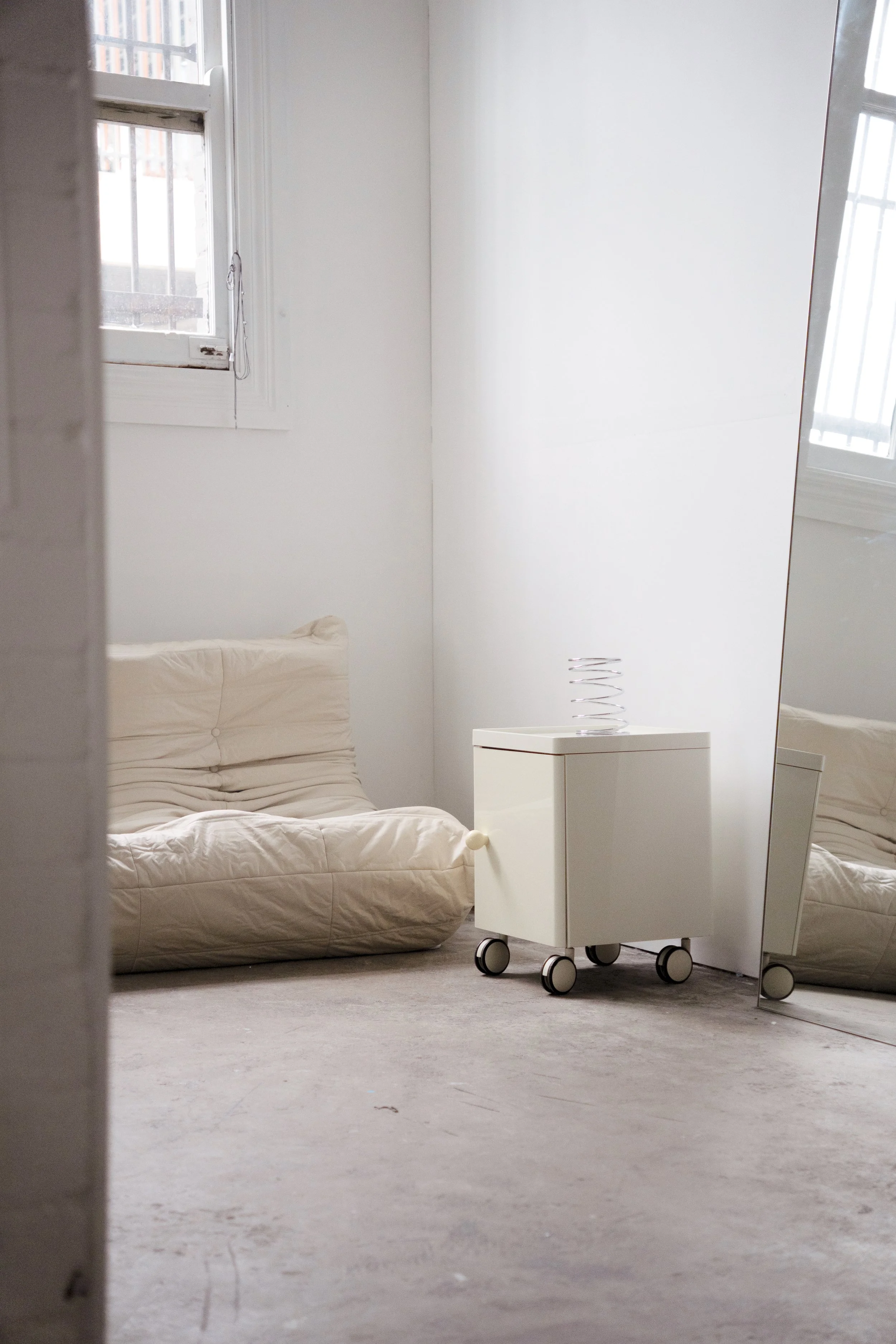 A minimalistic room with a beige futon on the left, a white side table on wheels with a spiral-shaped object on top, and a large mirror on the right corner. There are two small windows with white frames letting in natural light.