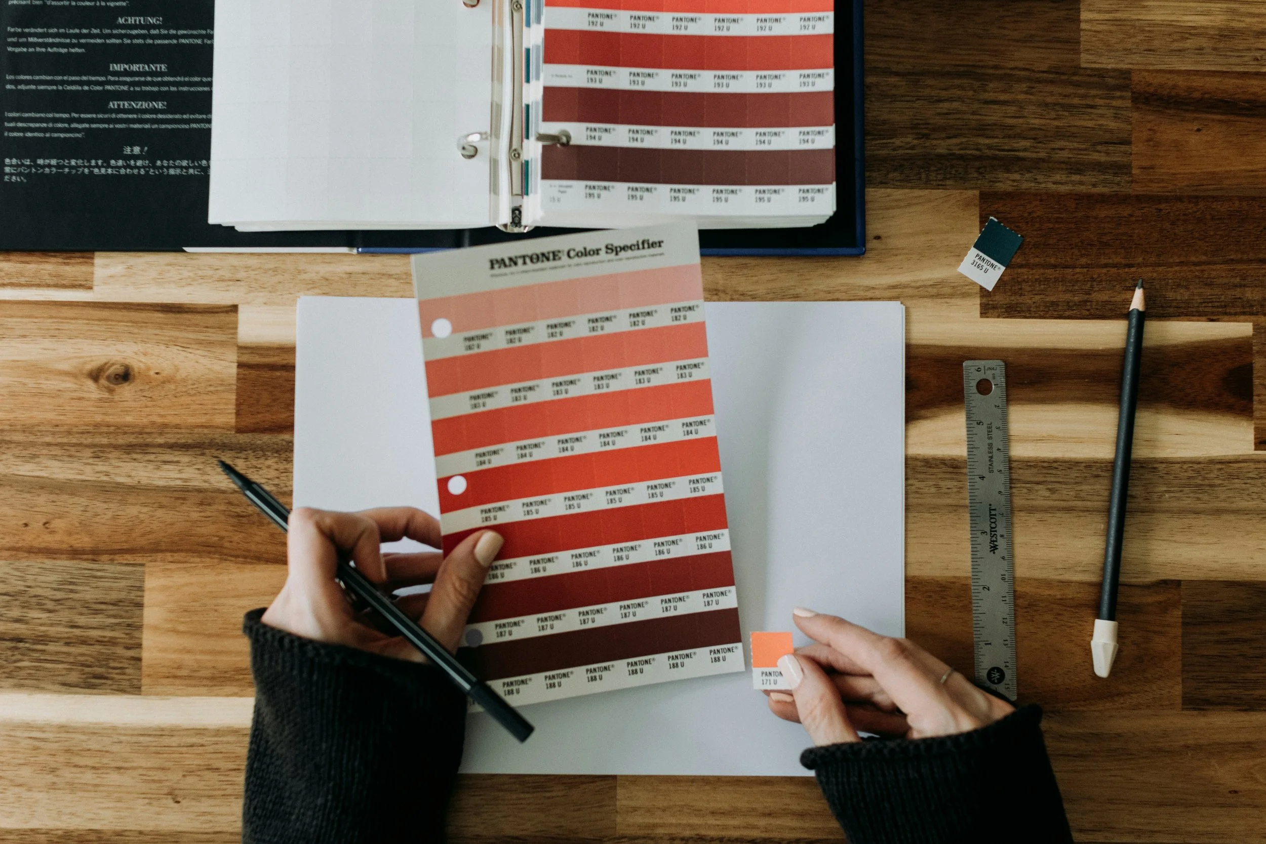 A person holds a Pantone color guide with various shades of red and orange, surrounded by a ruler, a pen, and a paint color sample on a wooden desk.