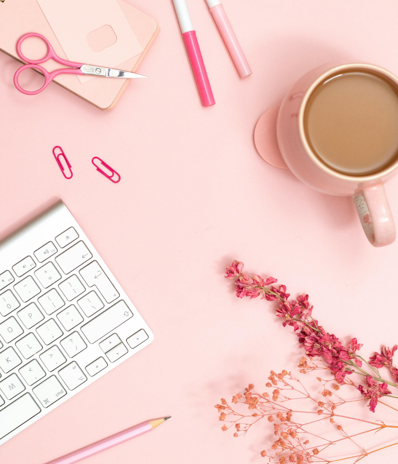 Flat lay of a pink desk with a white keyboard, pink scissors, pink paper clips, pink and white markers, a pink mug of coffee, a pink notepad, a pink pen, and dried pink and orange flowers.