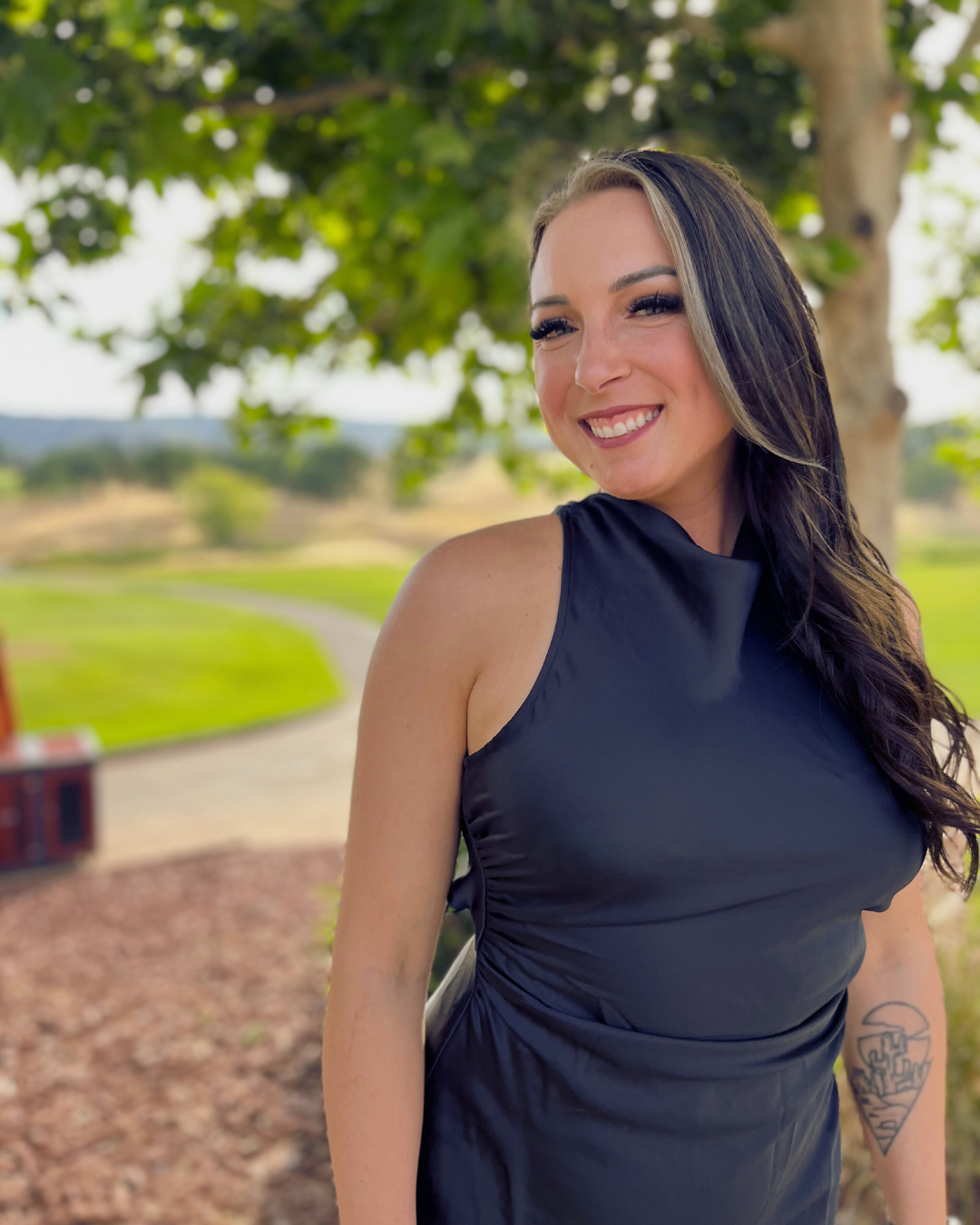 A smiling woman with long dark hair, wearing a black sleeveless dress, standing outdoors under a tree in a park.