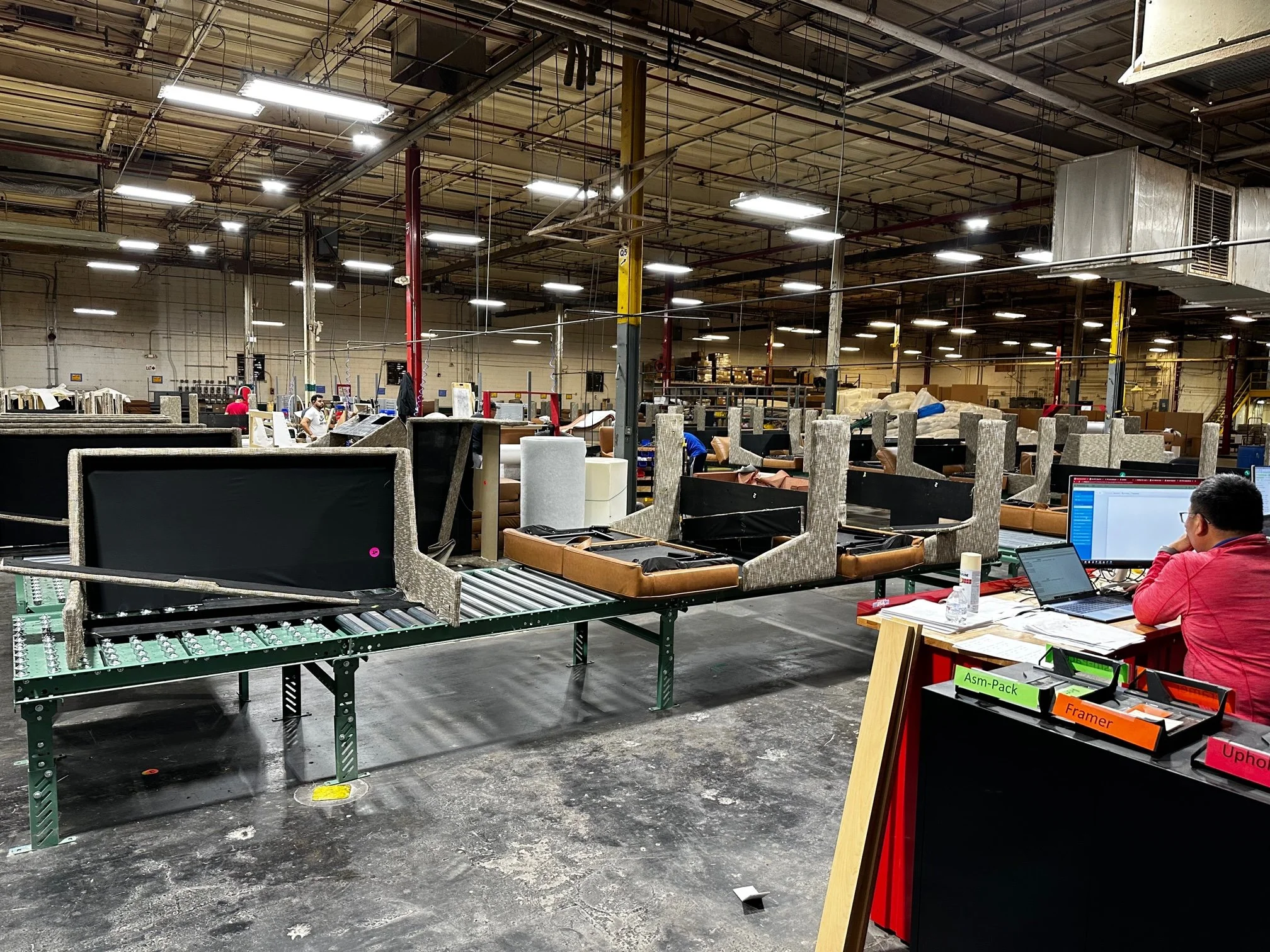 Warehouse with workers assembling furniture, conveyor belts, and a work station with computers and labeled bins.
