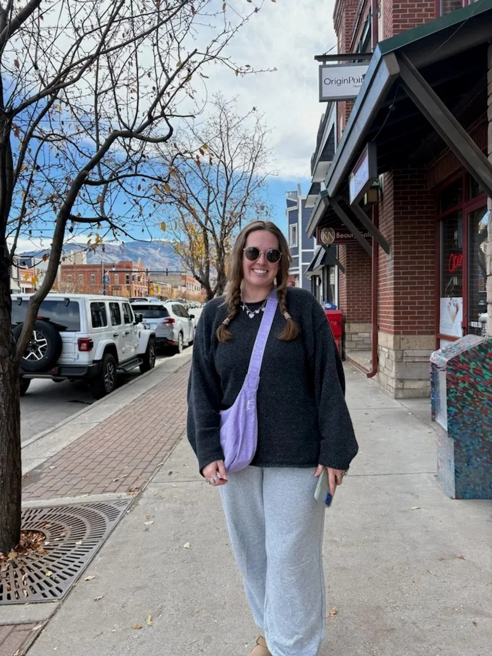 A smiling woman with long hair in braids, wearing sunglasses, a black sweater, and light gray sweatpants, standing on a city sidewalk next to a leafless tree. She has a purple shoulder bag and is holding a phone. There are parked cars, brick buildings, and mountains in the background, with a partly cloudy sky.