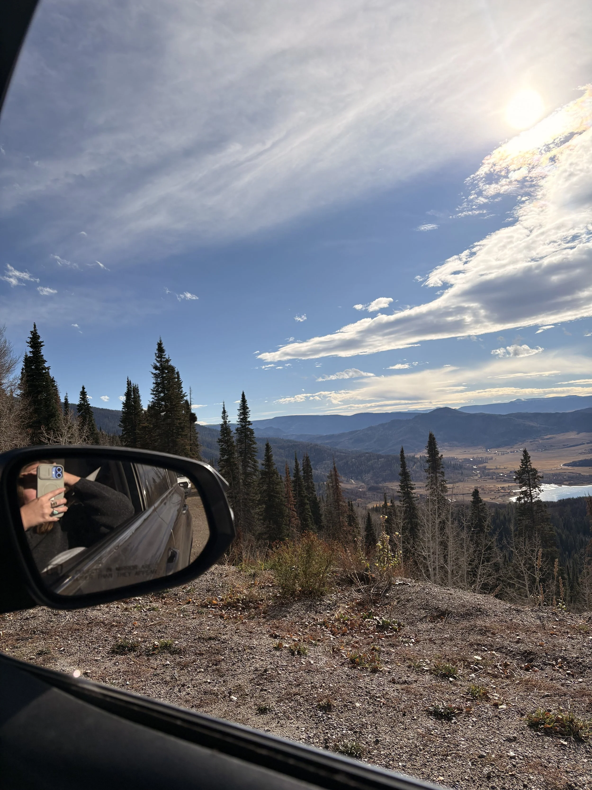 Scenic mountain landscape viewed from a car window with a side mirror in the foreground reflecting a person taking a selfie, under a partly cloudy sky with the sun shining.