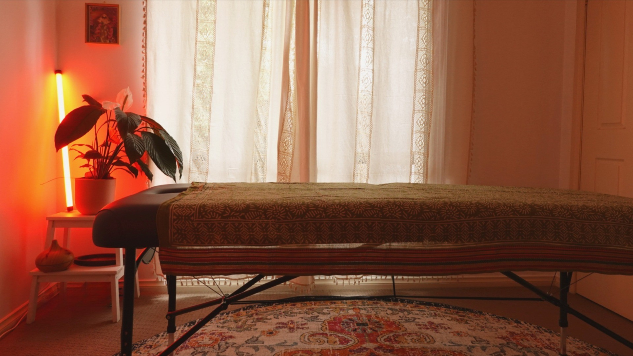 A massage table in a softly lit room with sheer curtains, a potted plant, a small side table, and a patterned rug.
