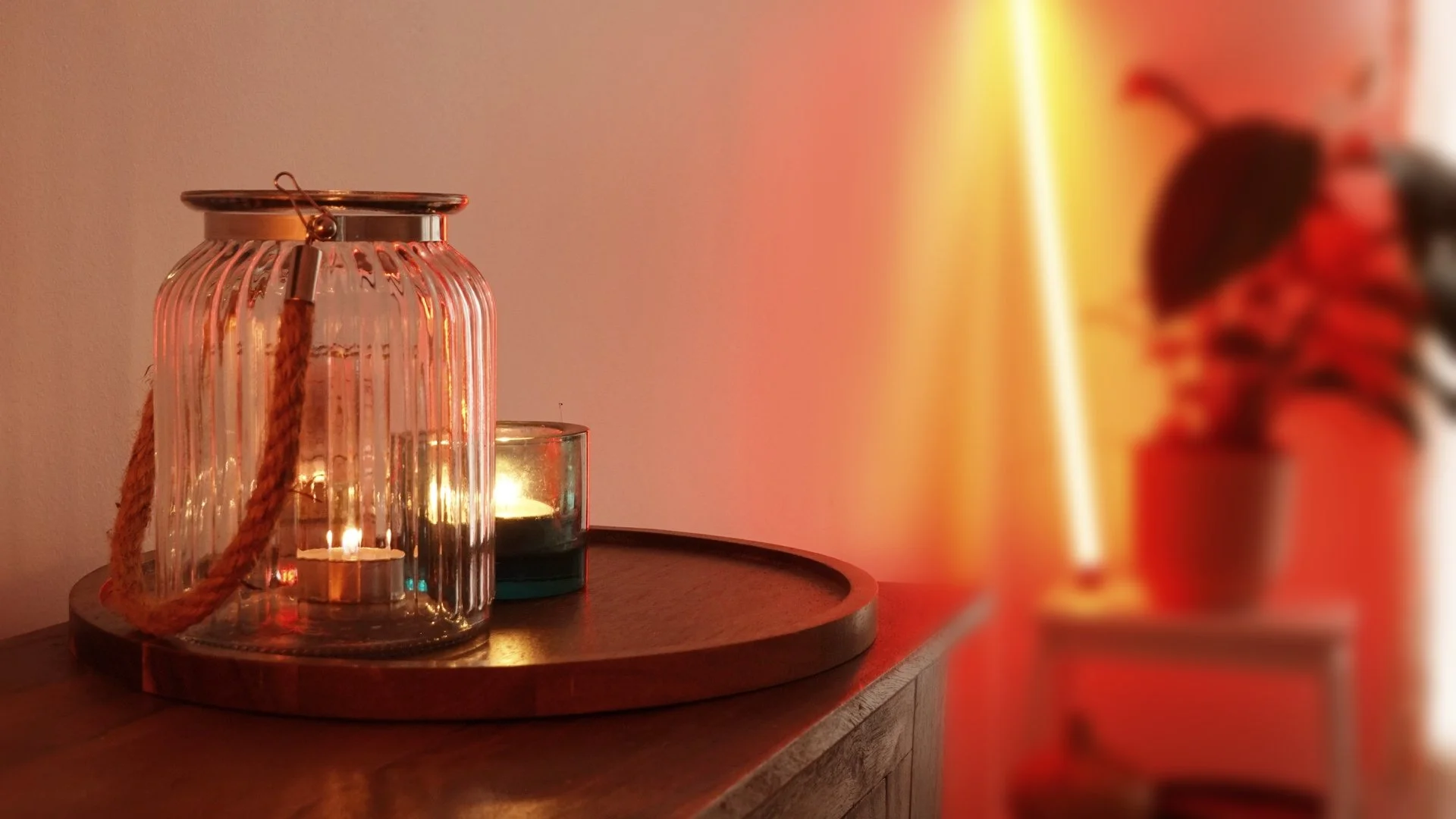 A decorative glass lantern with a lit candle inside, placed on a wooden surface with a round tray. In the background, there is a smaller tealight candle holder with a lit candle, and a blurred potted plant 's silhouette. Warm lighting creates a cozy atmosphere.