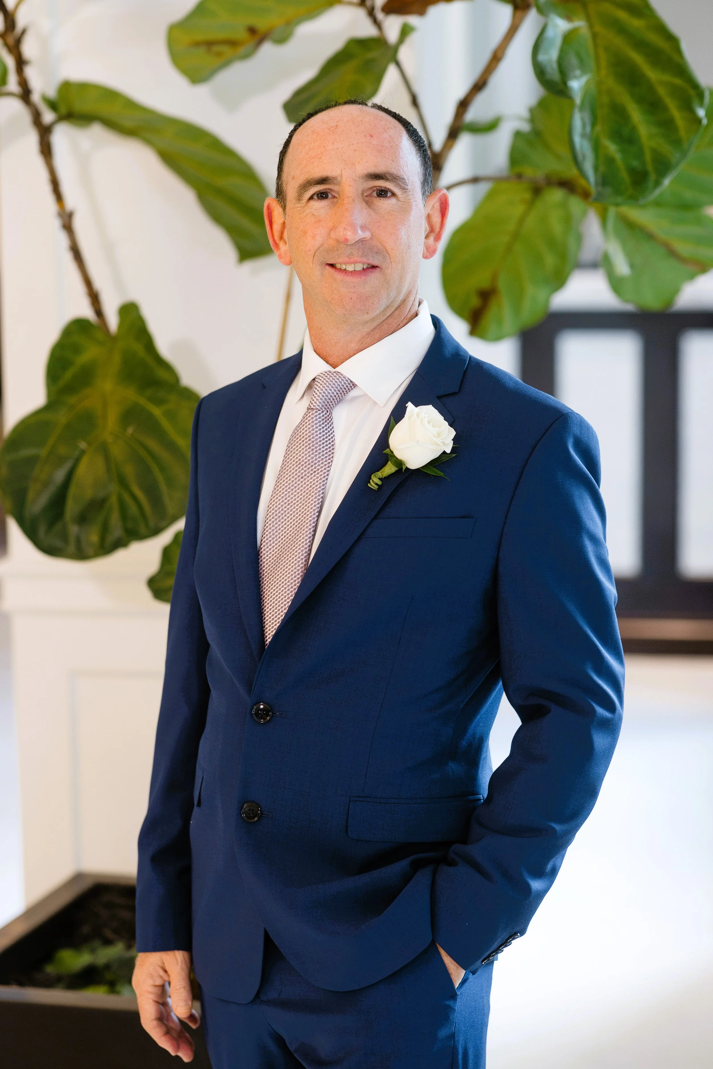 A man in a navy blue suit with a white shirt and a patterned tie, wearing a white rose boutonniere, standing indoors with a large green leafy plant in the background.
