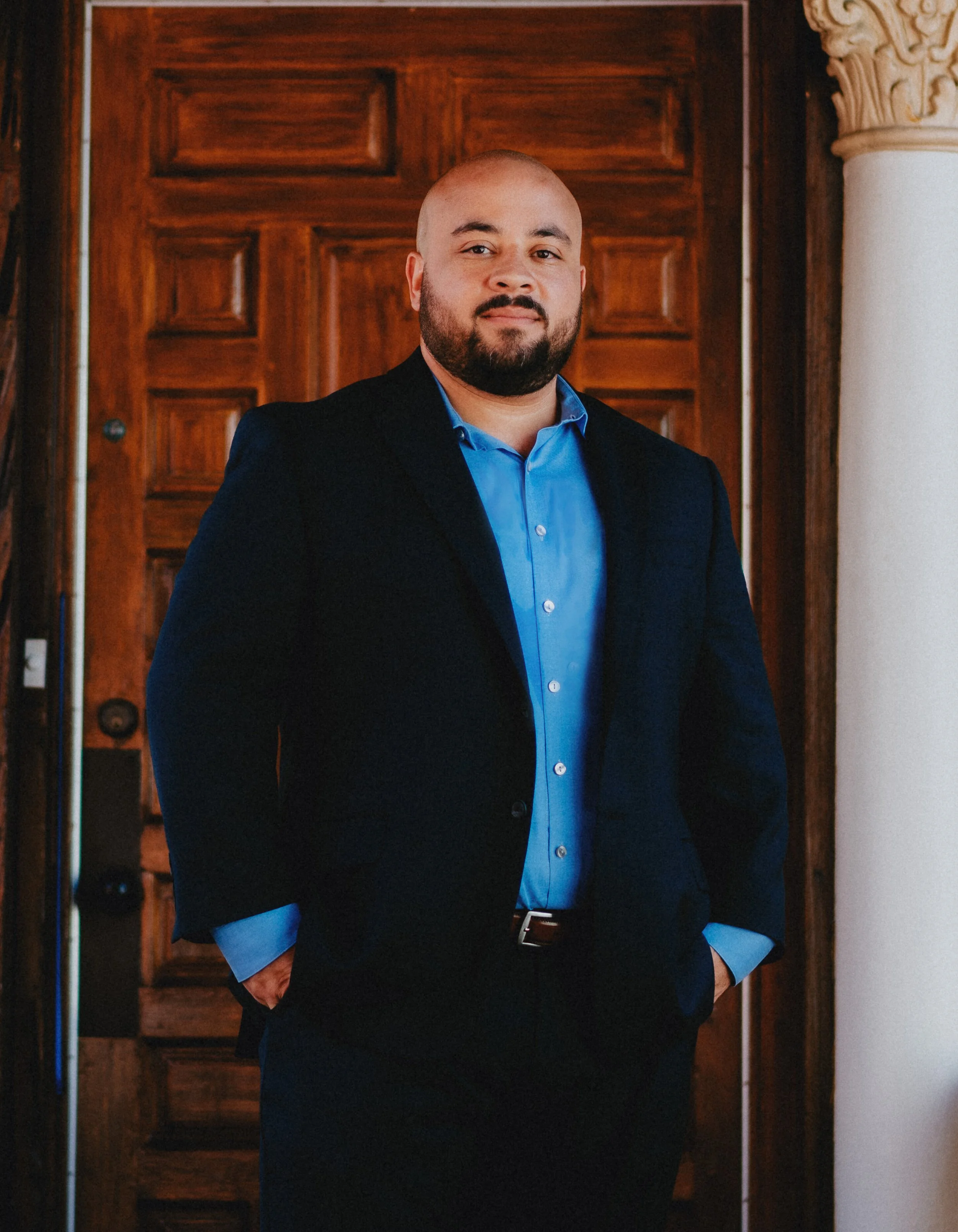 A man in a black suit and blue shirt standing indoors in front of a wooden door, with hands in his pockets, looking at the camera.