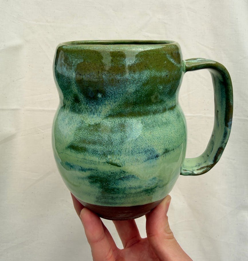 A green and brown ceramic mug with a handle, held by a person's hand against a light background.