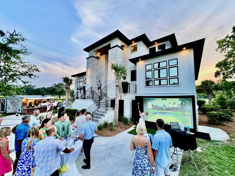 People gathered outside a modern house for a private party with a golf simulator being played.