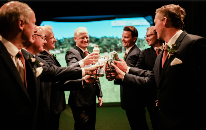 Wedding: Group of men in suits celebrating with drinks at a wedding or formal event, standing in front of a large golf simulator screen.