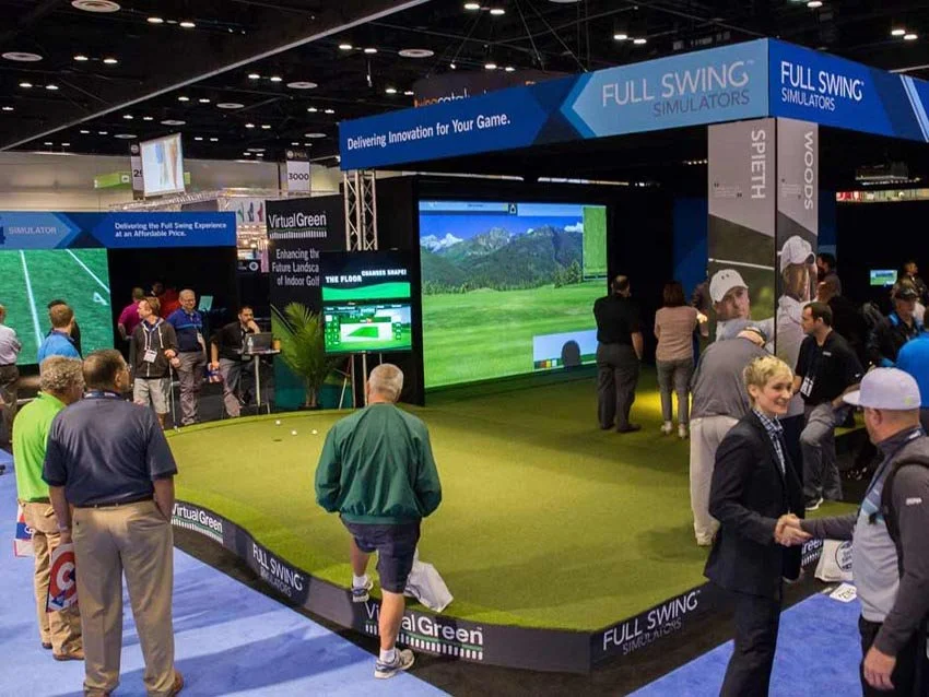 A golf simulator setup at a corporate event expo with a large screen displaying a golf course, surrounded by multiple people, including exhibitors and visitors, showcasing golf technology.