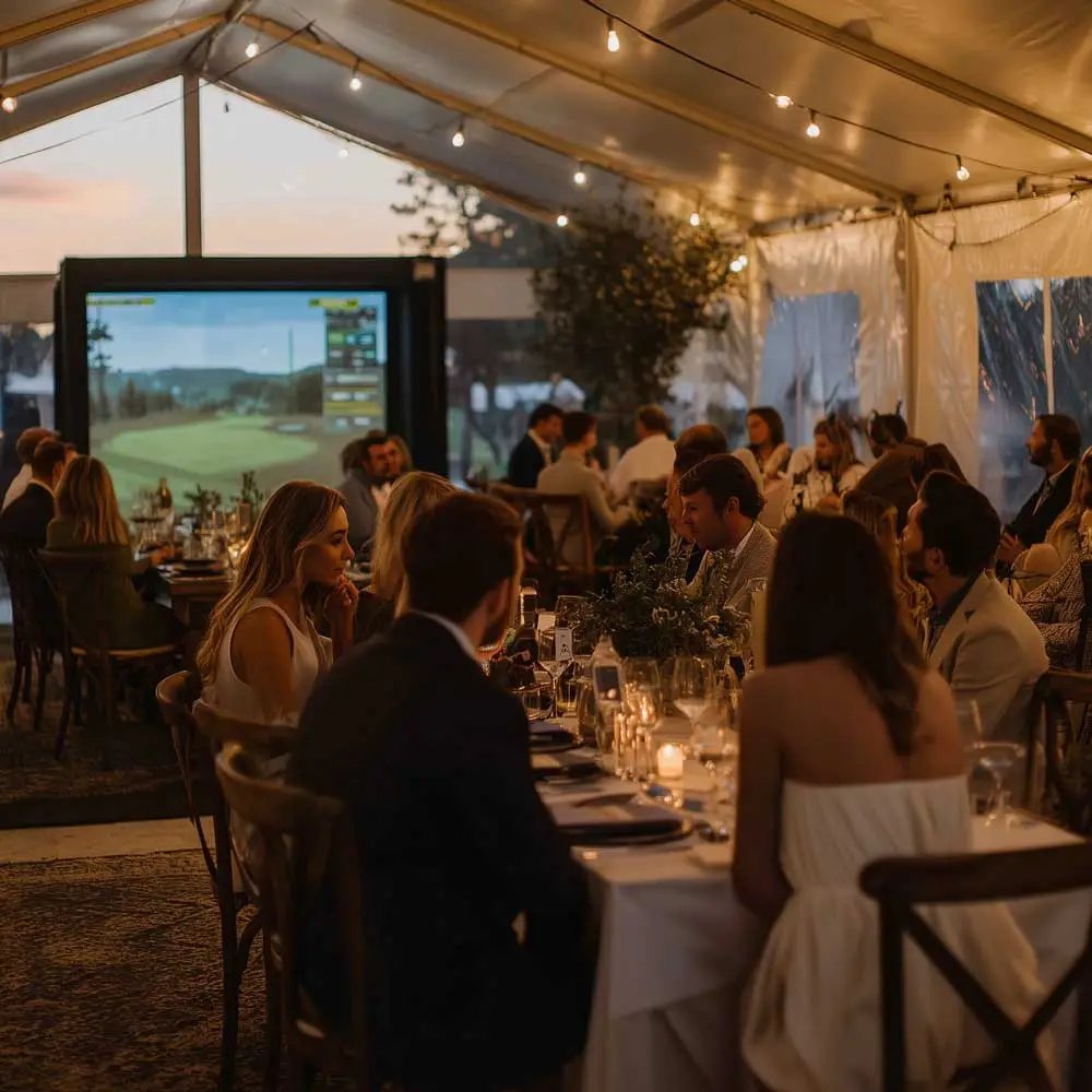 Wedding guests seated at a wedding dinner inside a tent with a golf simulator in the background.