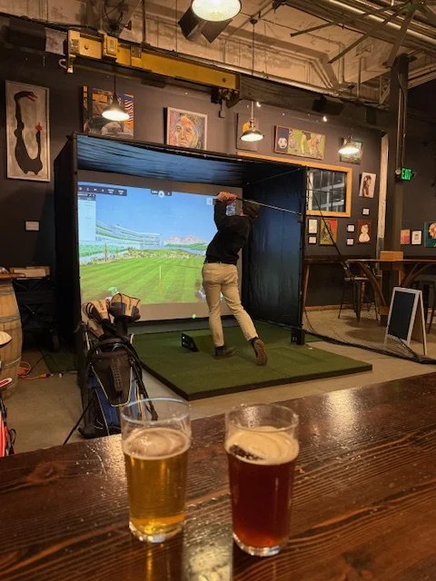 Person swinging a golf club on an indoor golf simulator, with two glasses of beer on the table in the foreground.