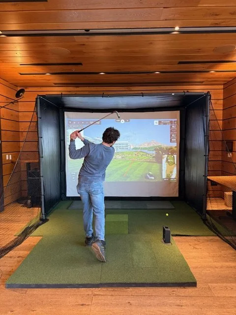 Indoor golf simulator with a person swinging a golf club, aiming at a large screen displaying a golf course, in a room with wooden walls and floor.