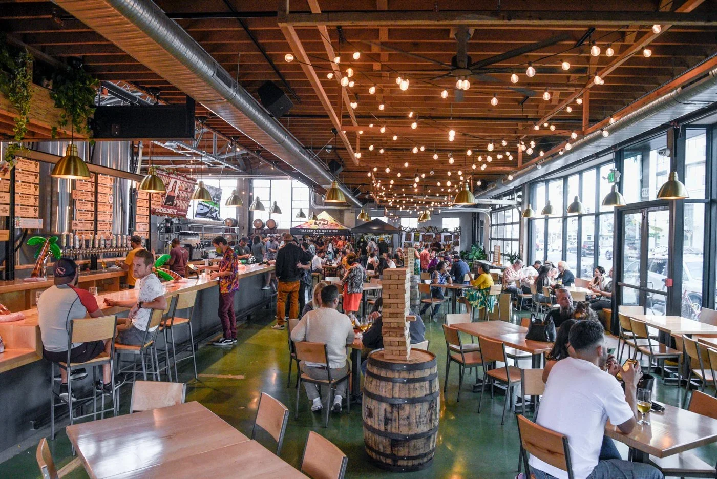 Retail Pop-Up: Interior of a busy modern brewery with large windows, wooden ceiling, string lights, bar counter on the left, and tables with patrons.
