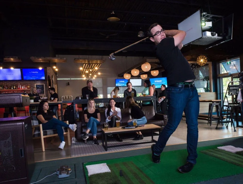 Person swinging a golf club on an indoor golf simulator with a group of people watching.
