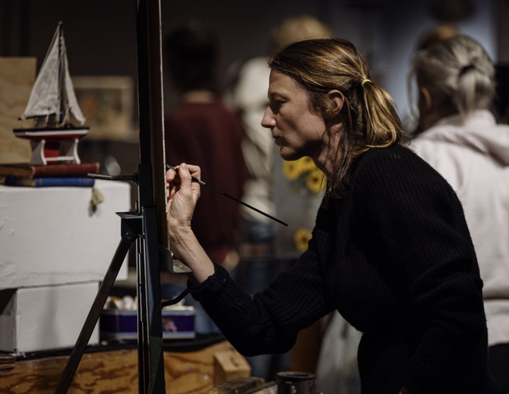 A woman painting on a canvas in an art studio, with other people in the background.