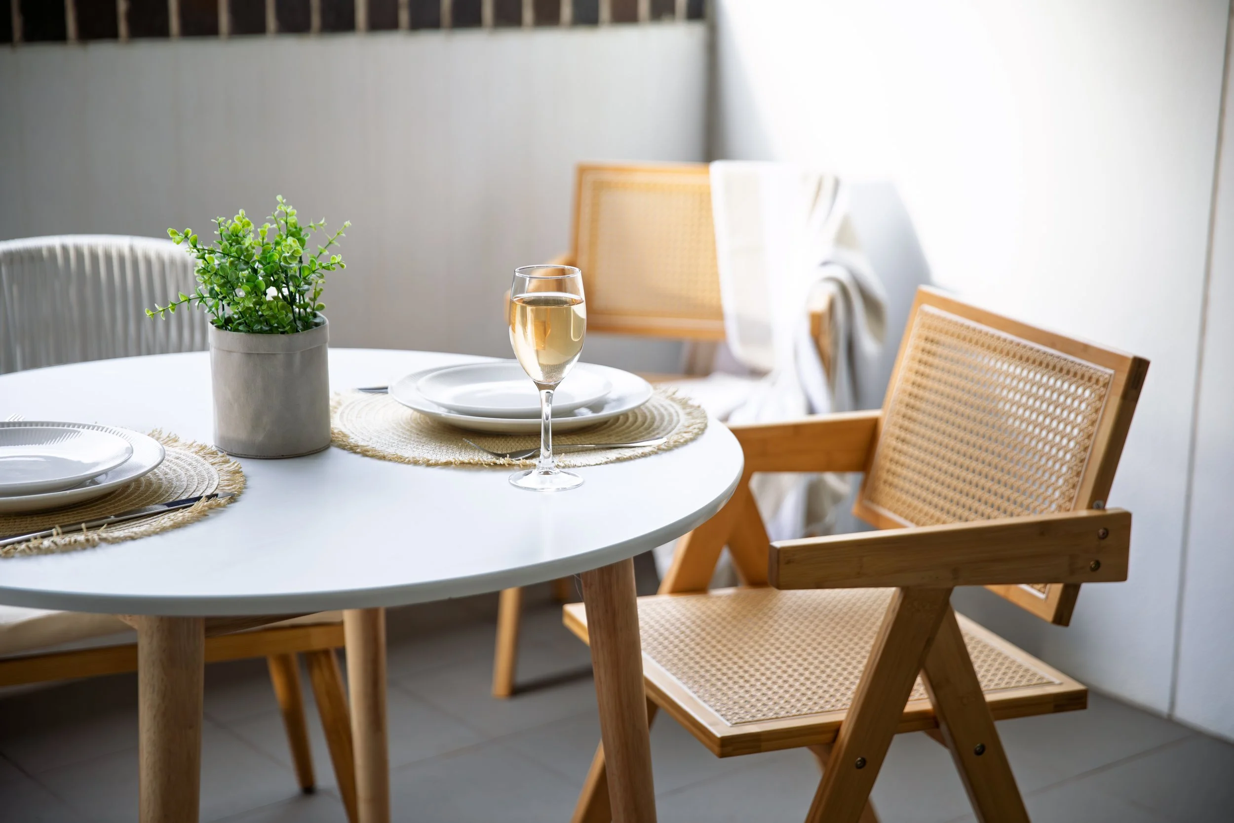 A round white dining table set with plates, napkins, and utensils, with a glass of white wine and a potted green plant at the center, surrounded by wooden and rattan chairs, in a bright room with natural light.