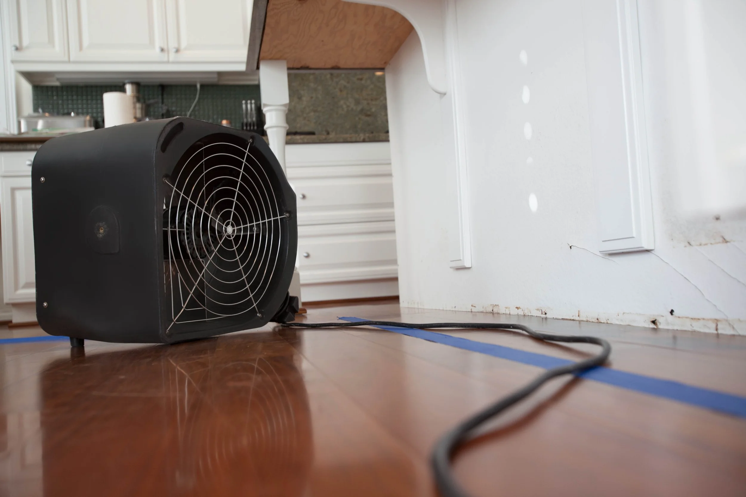 A black portable fan with a protective grill on the front, placed on a wood floor near a white wall with baseboards.