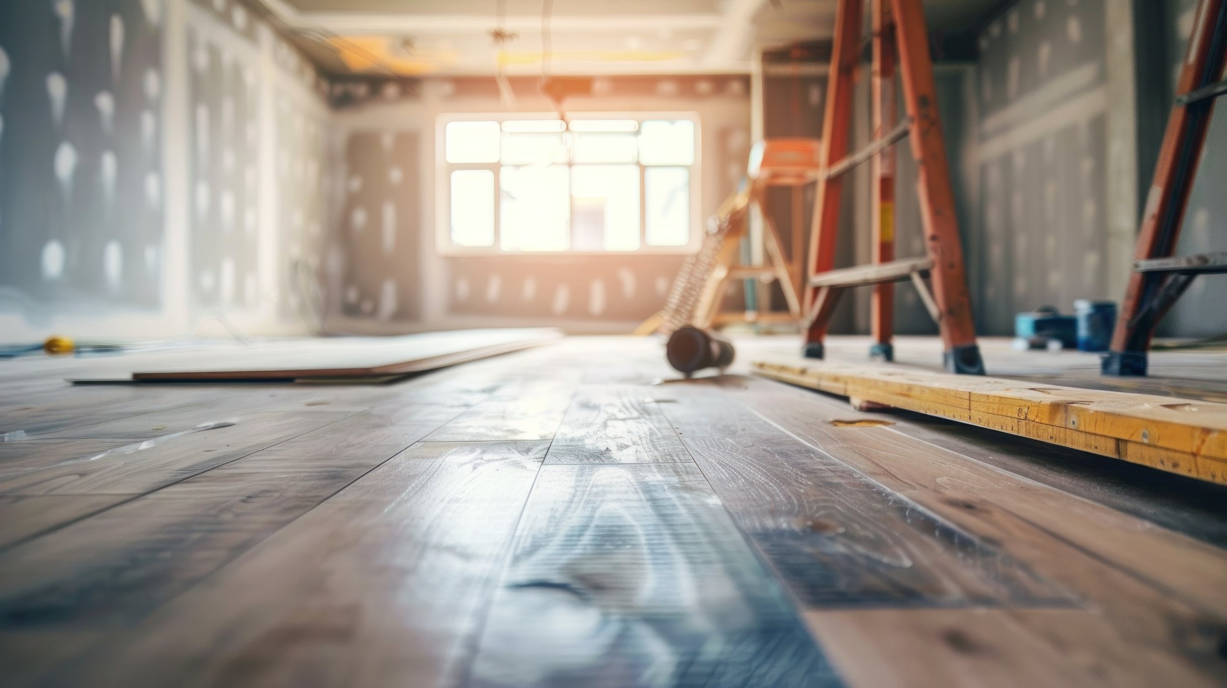 Interior of a room under construction with sunlight coming through a window, with tools, ladder, and building materials on the wooden floor.