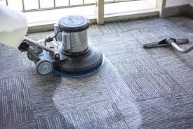 A floor cleaning machine used for carpet cleaning on a textured gray carpet, with a vacuum hose nearby, in front of a window.
