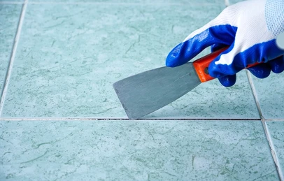 Someone in a blue and white glove using a trowel to apply grout between greenish tiles.