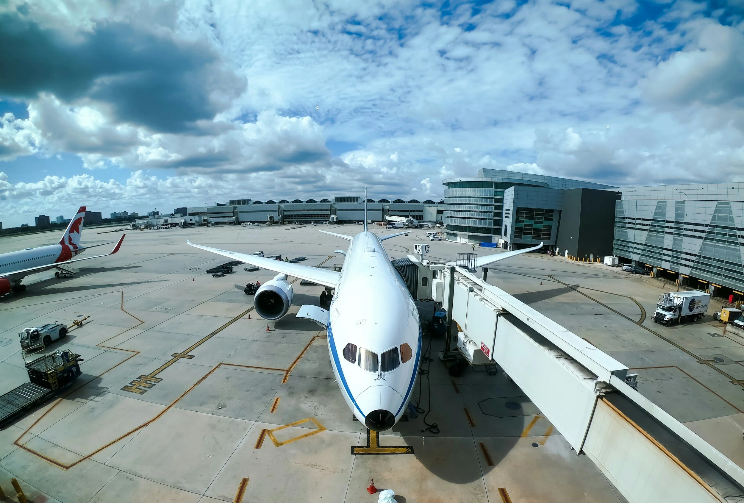 View of an airplane at an airport gate, with another plane to the left, and airport buildings and sky with clouds in the background.