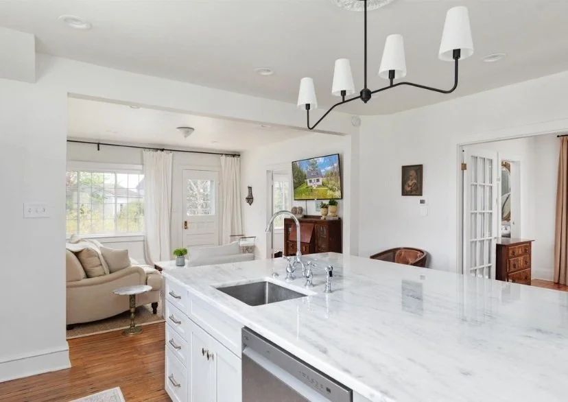 Interior view of a modern kitchen with a white marble island, a sink, and a hanging light fixture, looking into a living room with a beige sofa and a large window.
