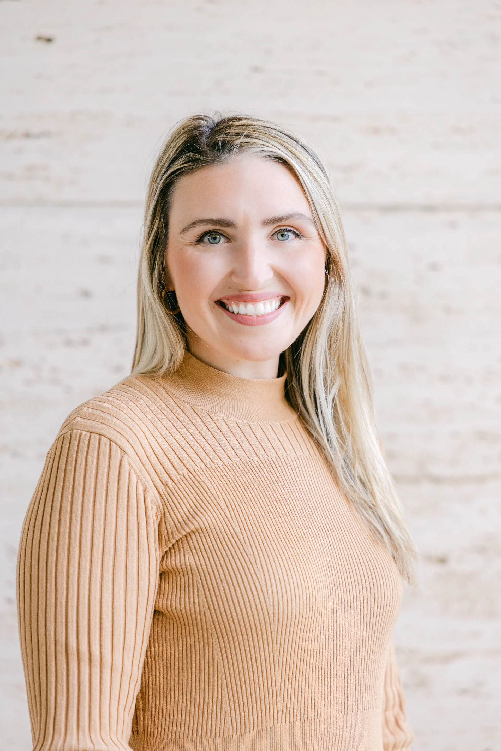 A woman with blonde hair smiling and wearing a beige ribbed sweater, standing against a light-colored brick wall.