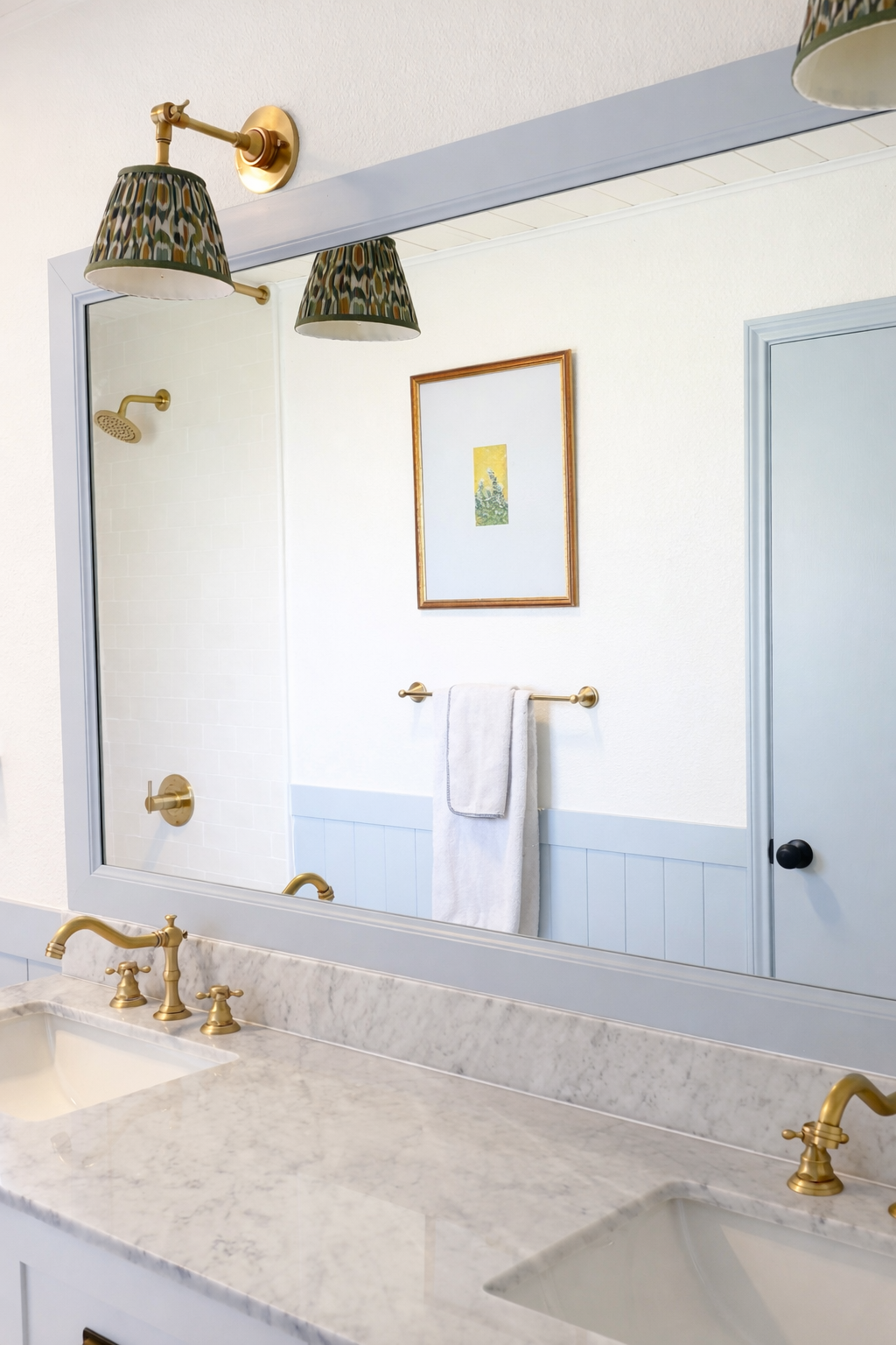 A bathroom with a marble countertop and gold fixtures, including a double sink, seen through a large mirror. The wall features a framed artwork and a towel hanging on a gold towel bar. There is a shower area with a gold showerhead in the background a