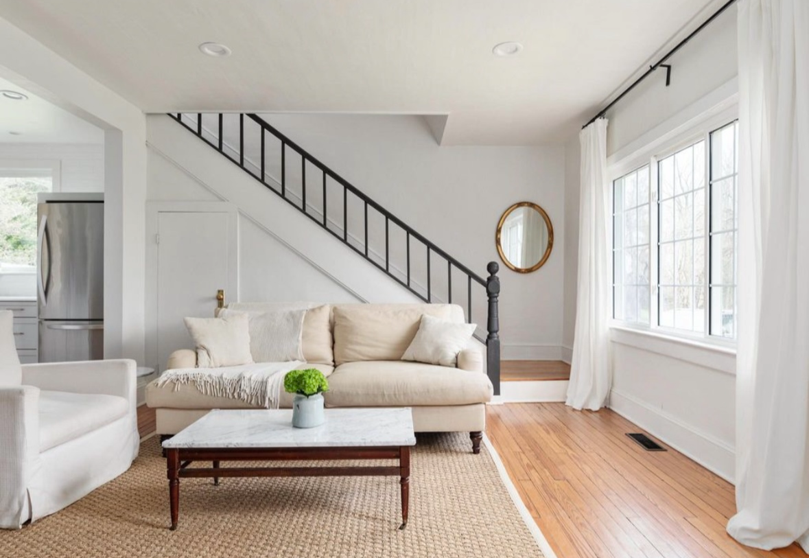 Bright living room with large windows, white curtains, cream sofa with throw pillows, coffee table with a green plant, beige rug, wooden floor, staircase with black railing, and a mirror on the wall.