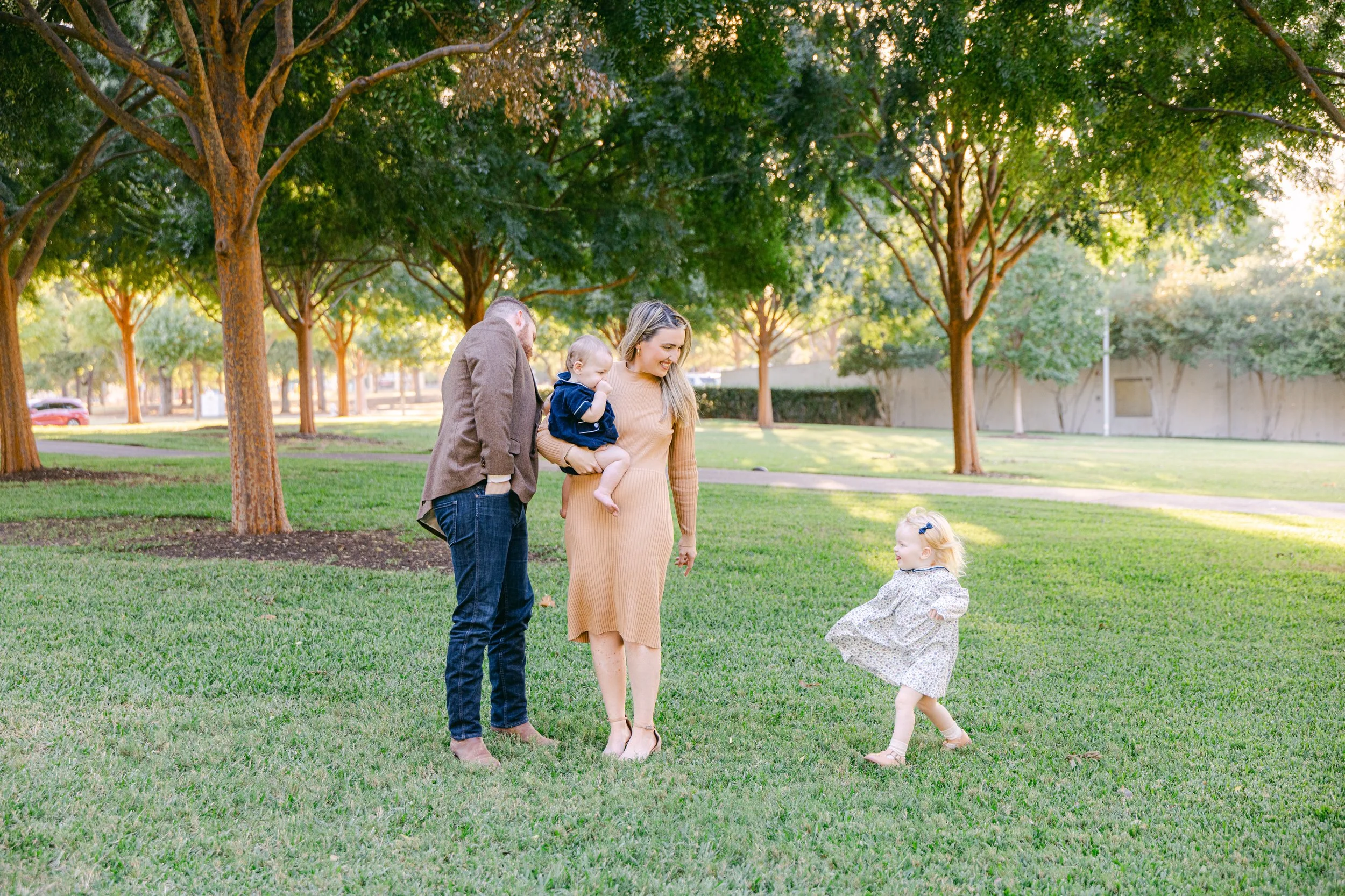 A family of four, including a man, woman, a young girl, and a toddler boy, enjoying time in a park with lush green grass and tall trees. The woman is holding the toddler, and the young girl in a dress is happily dancing or playing on the grass.